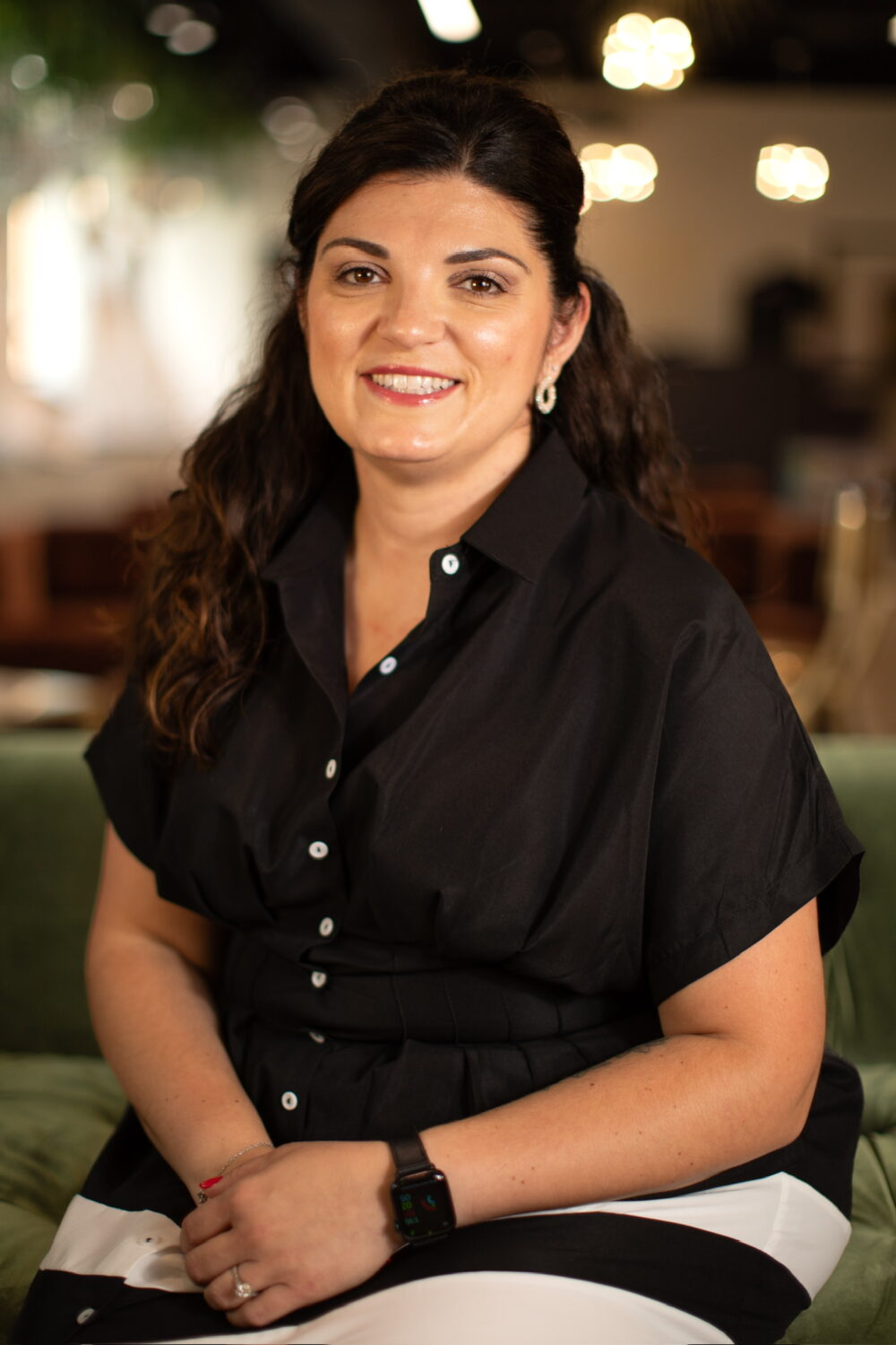 Smiling woman with dark shoulder-length hair in a black blouse, seated against a softly blurred, warmly lit indoor background.