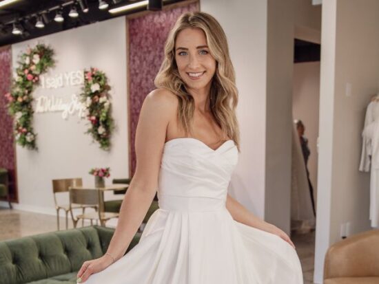 A bride in a strapless a-line wedding dress poses inside a bridal shop holding the skirt out to the sides.