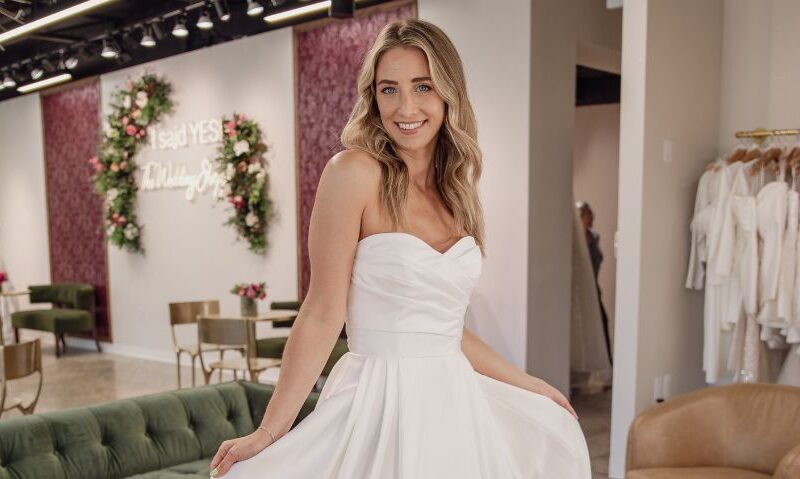 A bride in a strapless a-line wedding dress poses inside a bridal shop holding the skirt out to the sides.