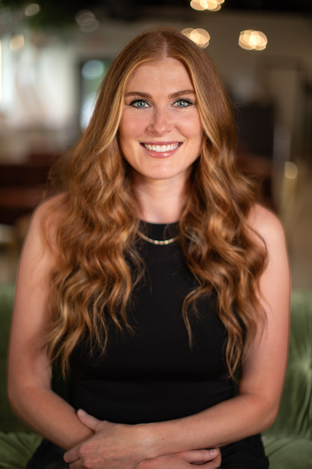 Smiling woman with long wavy red hair wearing a sleeveless black top, seated in a warm, softly blurred indoor setting