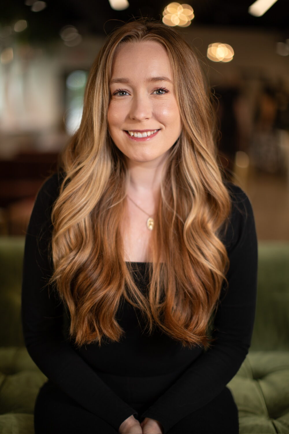 Smiling woman with long wavy blonde hair in a black long-sleeve top, seated against a softly lit, blurred indoor background
