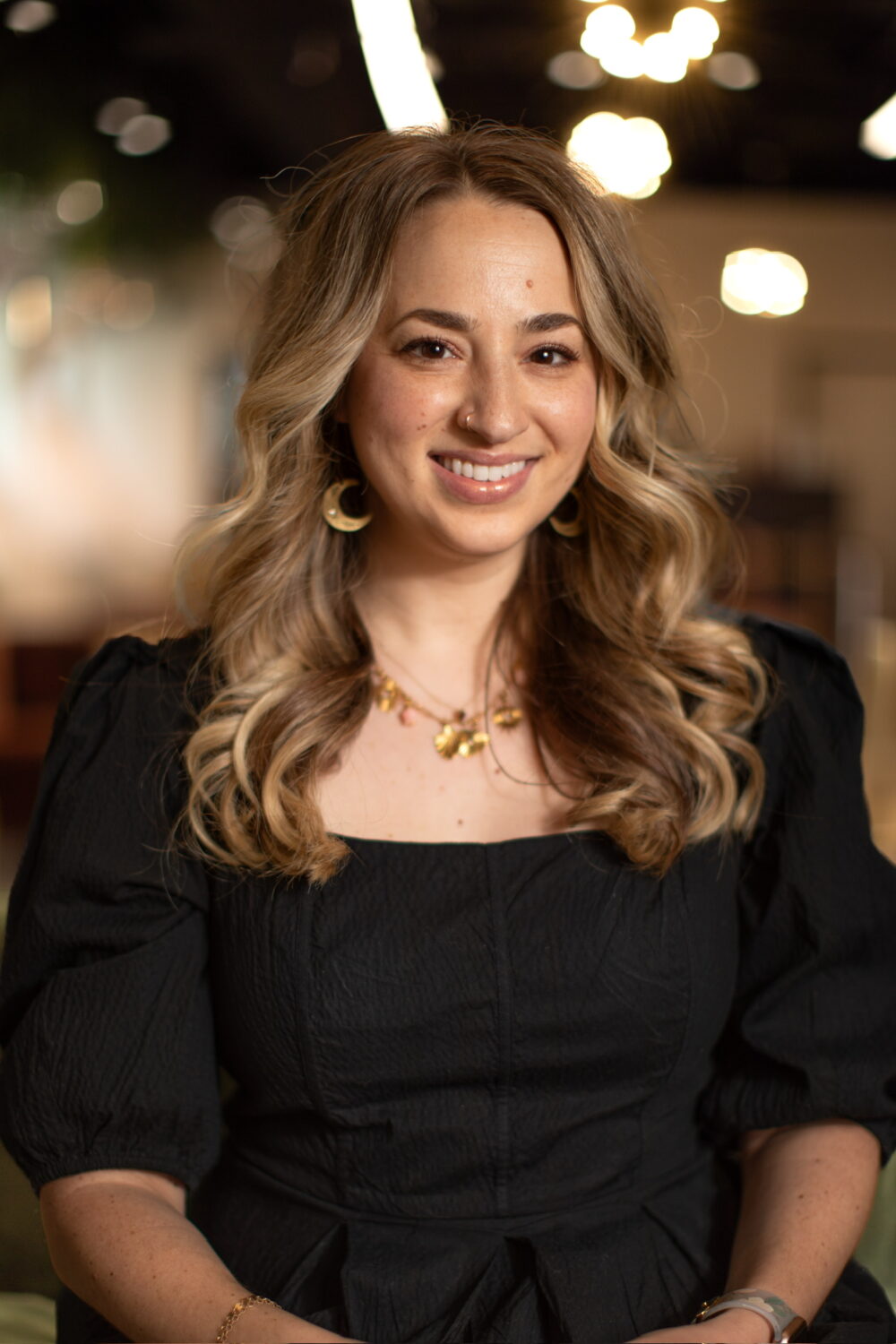 Smiling woman with long light brown wavy hair wearing a black dress and gold jewelry, seated in a warmly lit indoor space.