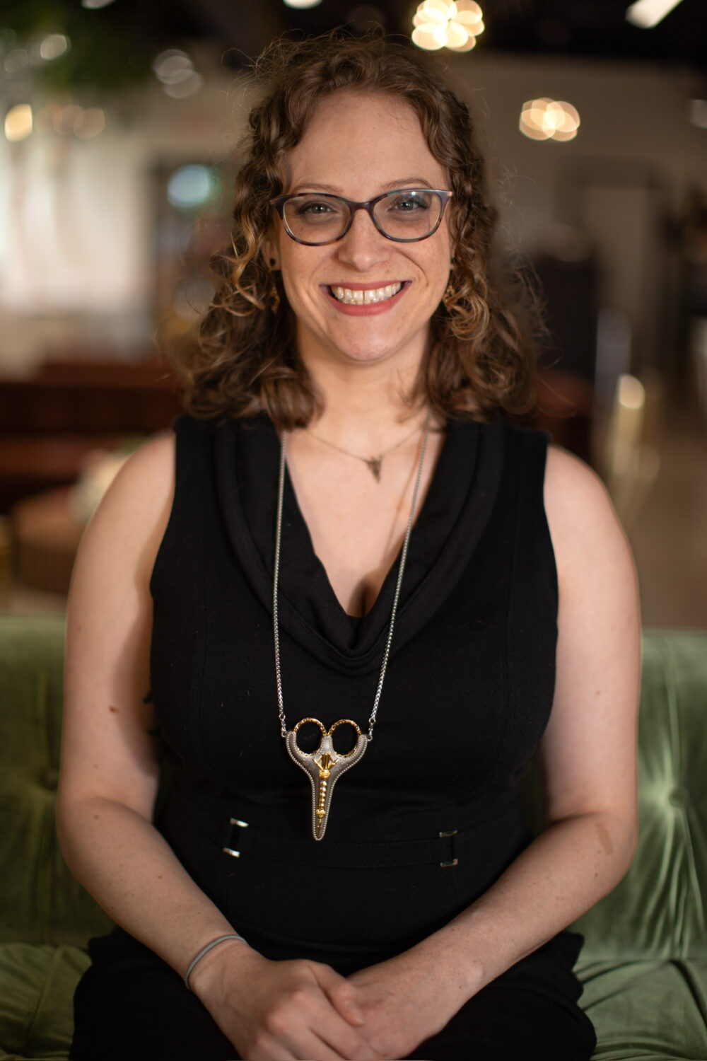 Smiling woman with curly light brown hair and glasses wearing a sleeveless black top and statement necklace, seated indoors with warm lighting.