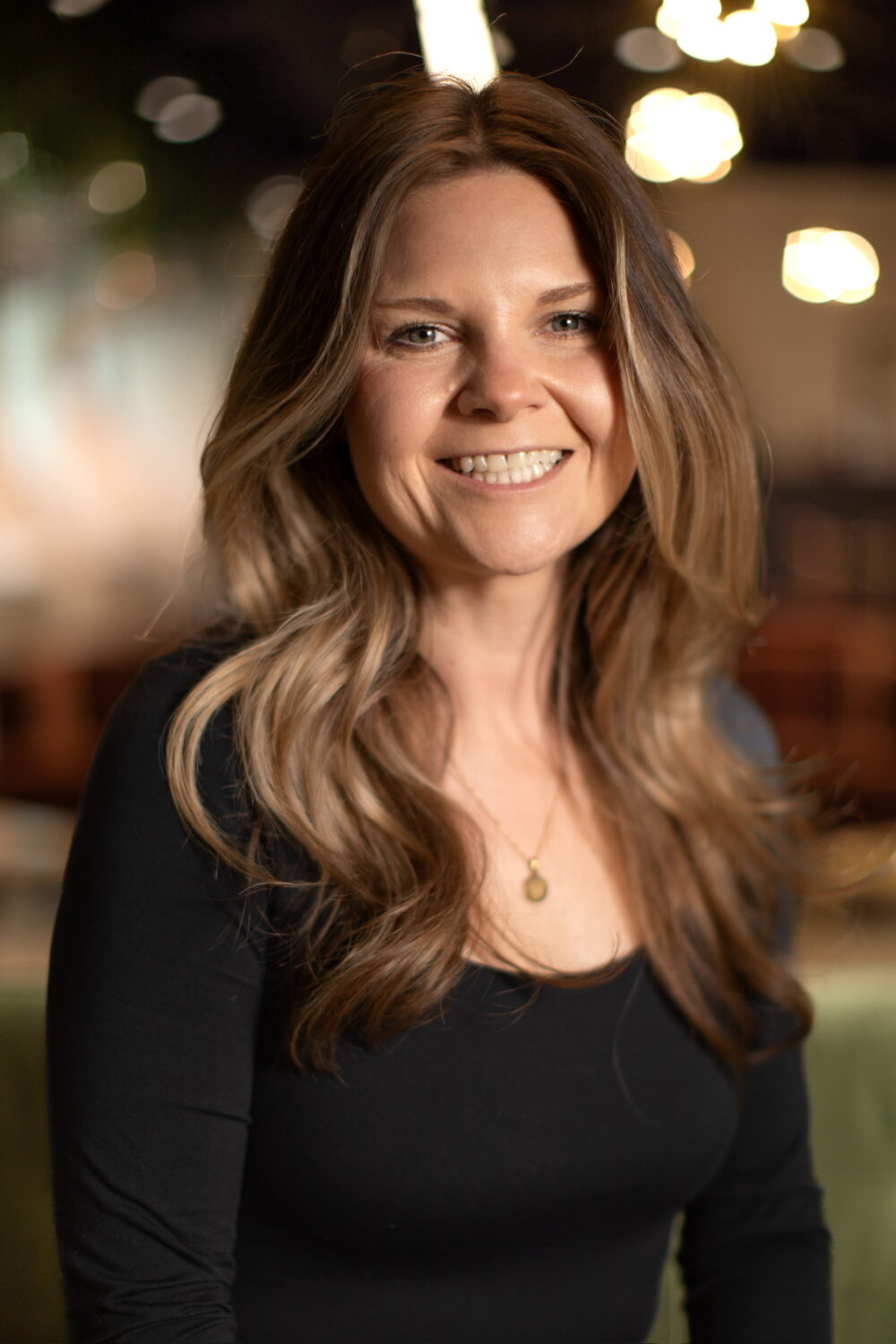 Smiling woman with long light brown hair wearing a black top, photographed in a softly blurred, warmly lit indoor setting.