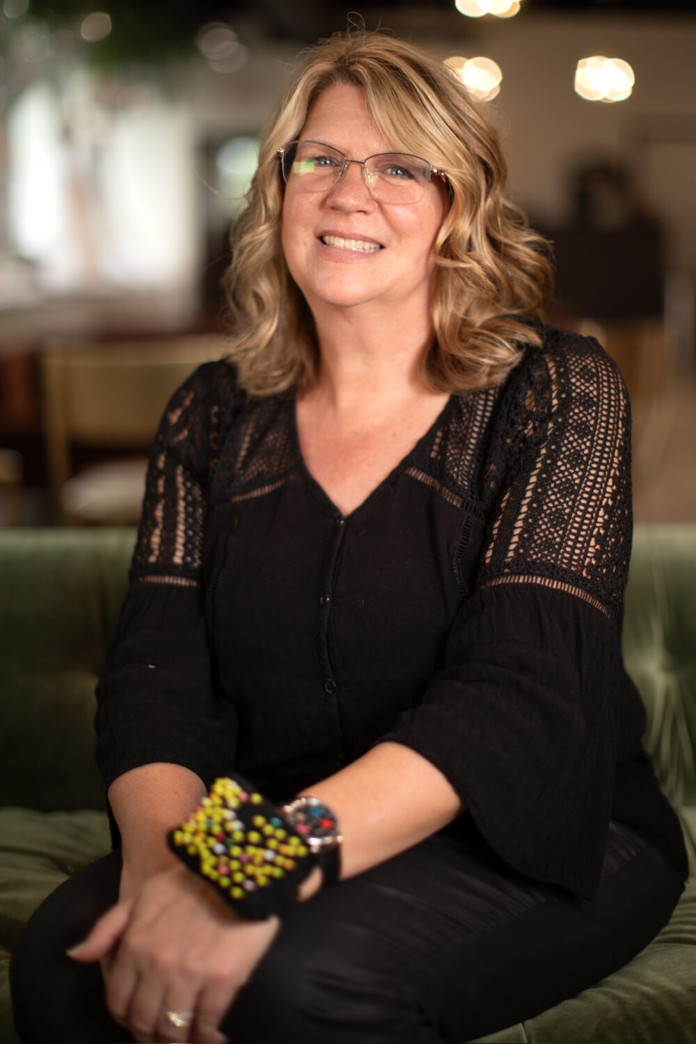 Smiling woman with shoulder-length blonde hair and glasses wearing a black outfit, seated in a warm, softly blurred indoor environment.