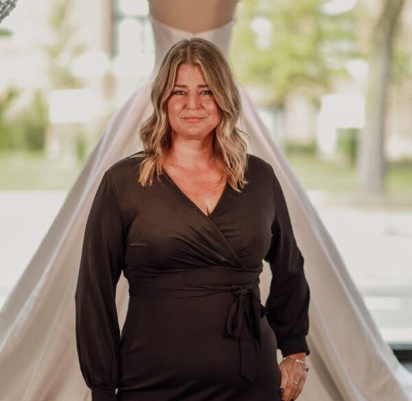 Smiling woman with shoulder-length blonde hair wearing a black dress, standing behind a wedding dress on display.