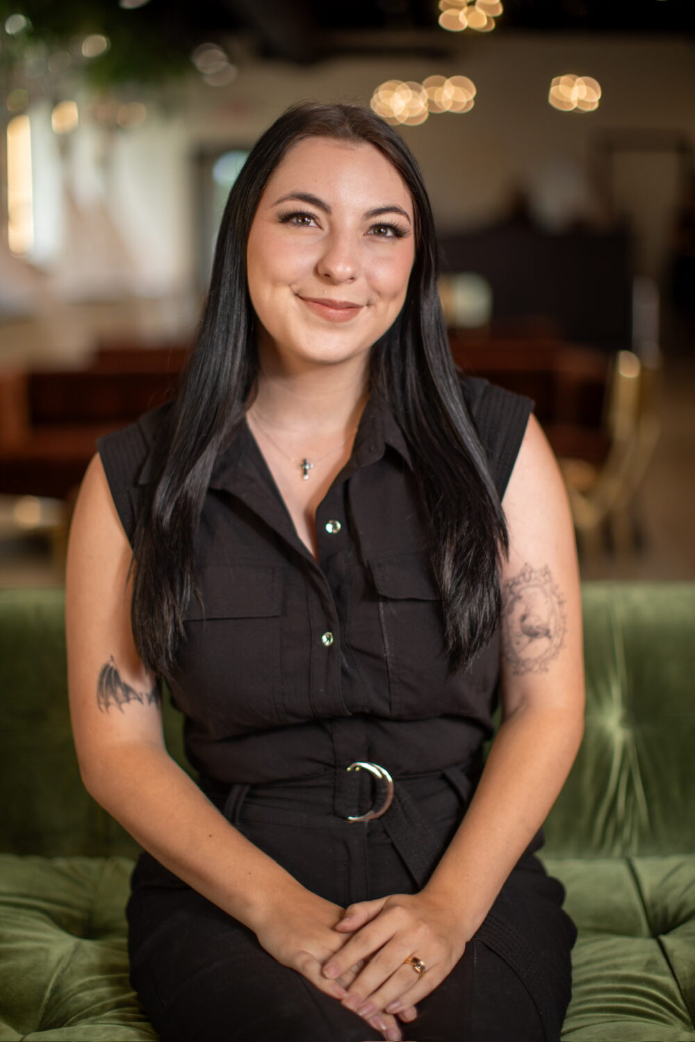 Smiling woman with long dark hair wearing a sleeveless black outfit, seated with visible arm tattoos in a softly lit indoor space.