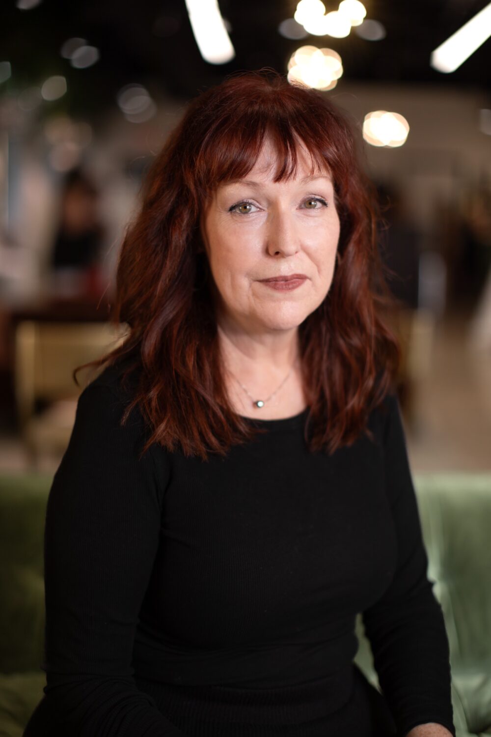 Smiling woman with red hair wearing a black top, photographed against a warm, softly blurred indoor background.