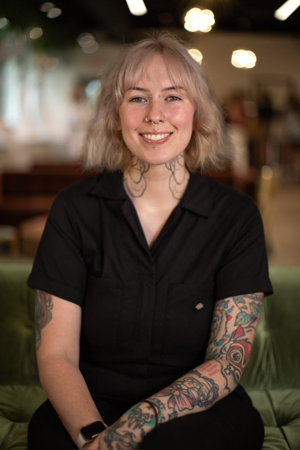 Smiling woman with short blonde hair and visible arm tattoos wearing a black collared shirt, seated in a warmly lit indoor setting.