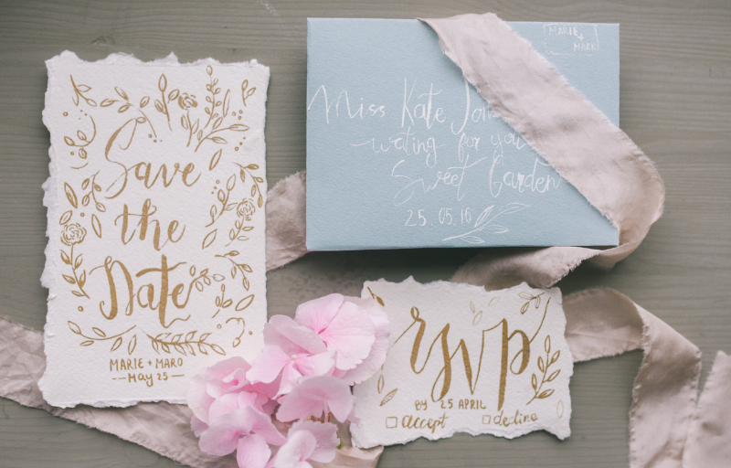 A flat-lay display of a wedding invitation suite including save-the-date cards, envelopes, and a ribbon arranged with pink flowers.