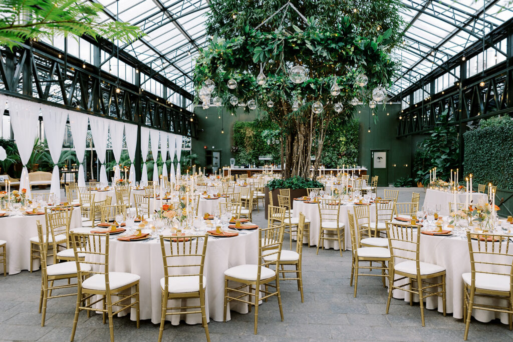 An open greenhouse filled with round tables set with gold chairs, white linens, and elegant centerpieces surrounded by lush greenery.