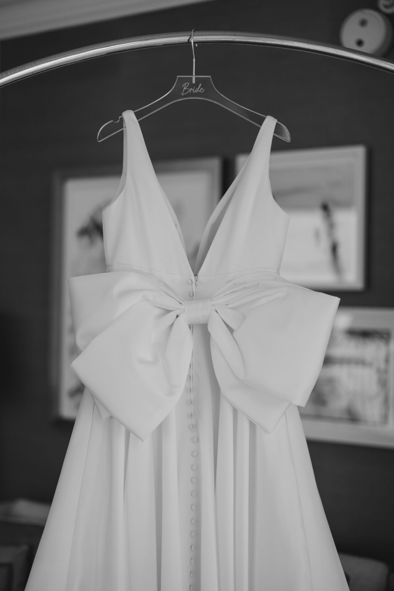 A white wedding dress with a large bow detail hangs from a hanger on a doorway inside a room.