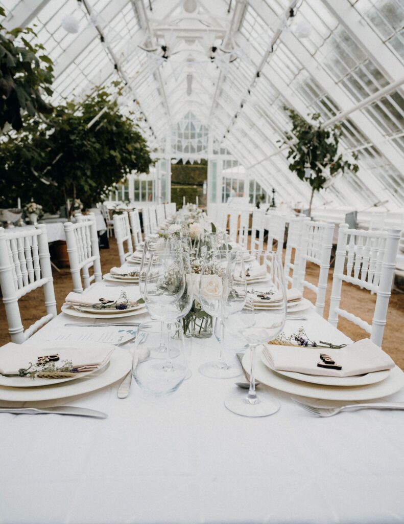 A long reception table inside a greenhouse is set with white linens, greenery centerpieces, and elegant table settings.