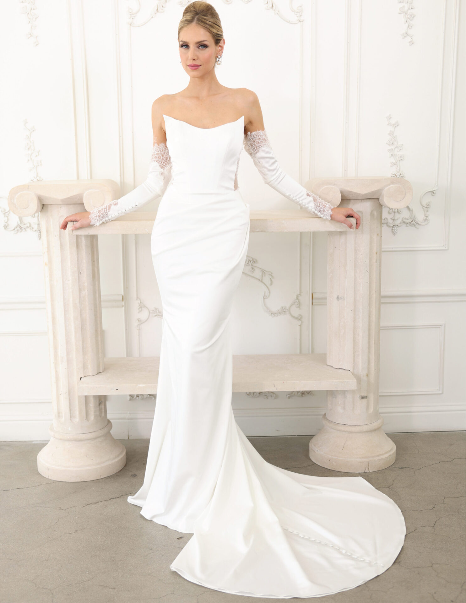 Bride in a strapless white couture gown with sheer sleeves, posed in an elegant white studio.
