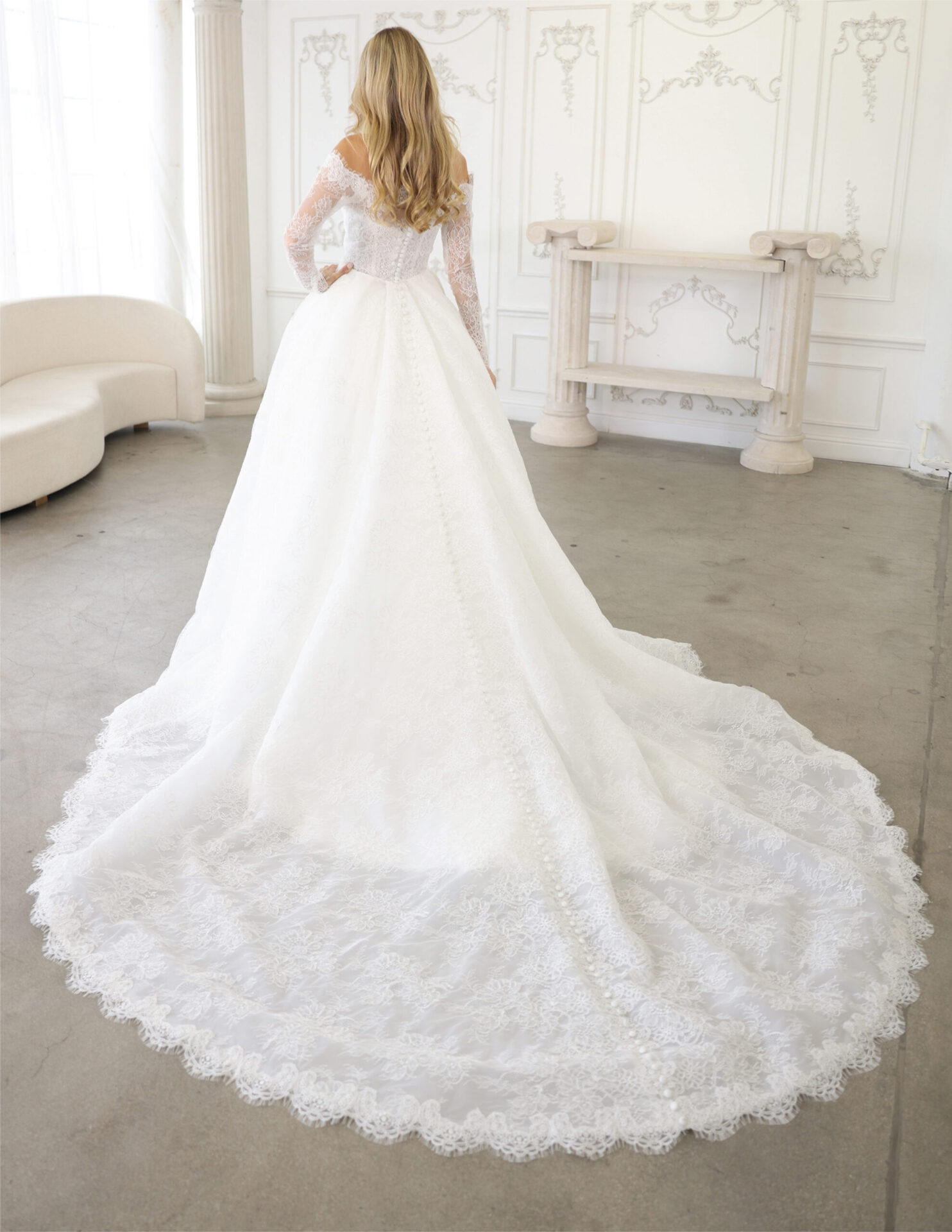 Back view of a bride in an off-the-shoulder lace couture gown with a circular train in a bright studio.