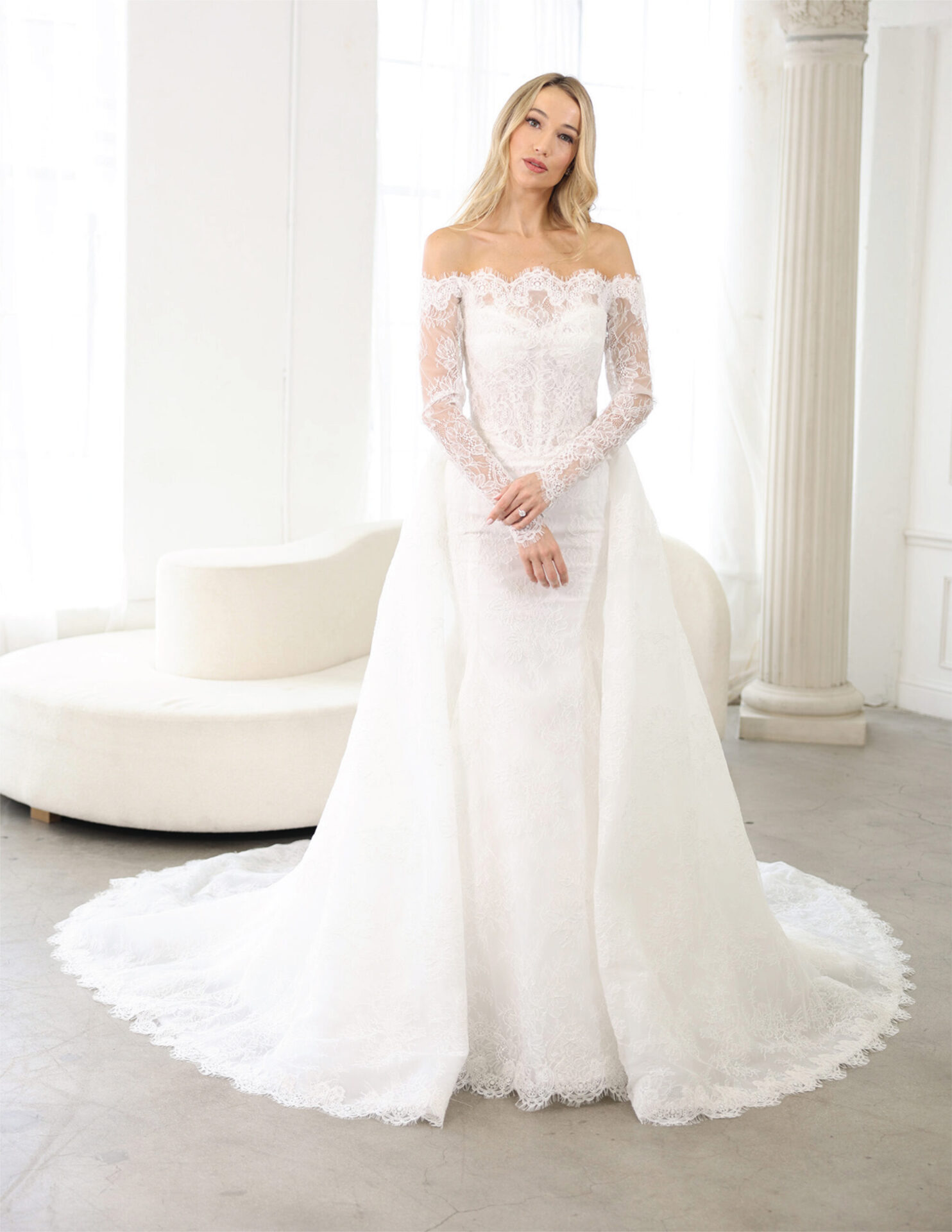 Bride in an off-the-shoulder lace wedding gown standing in a sunlit white studio.
