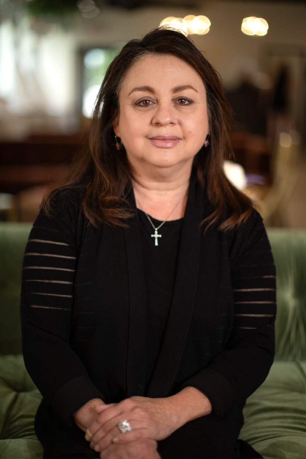 Woman with dark shoulder-length hair wearing a black top and cross necklace, seated against a softly blurred indoor background.