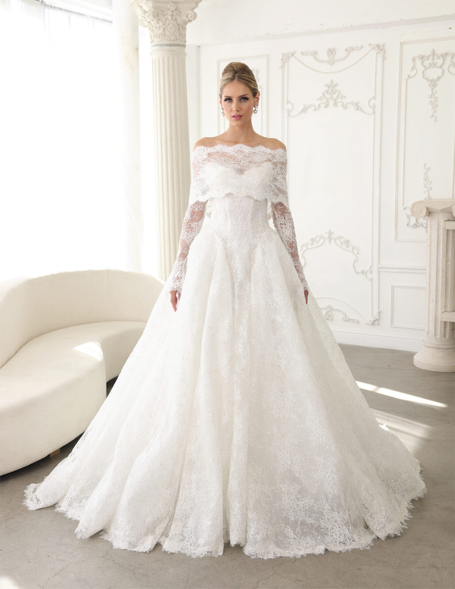Bride in an off-the-shoulder lace ball gown with a full skirt posed in an elegant white room.