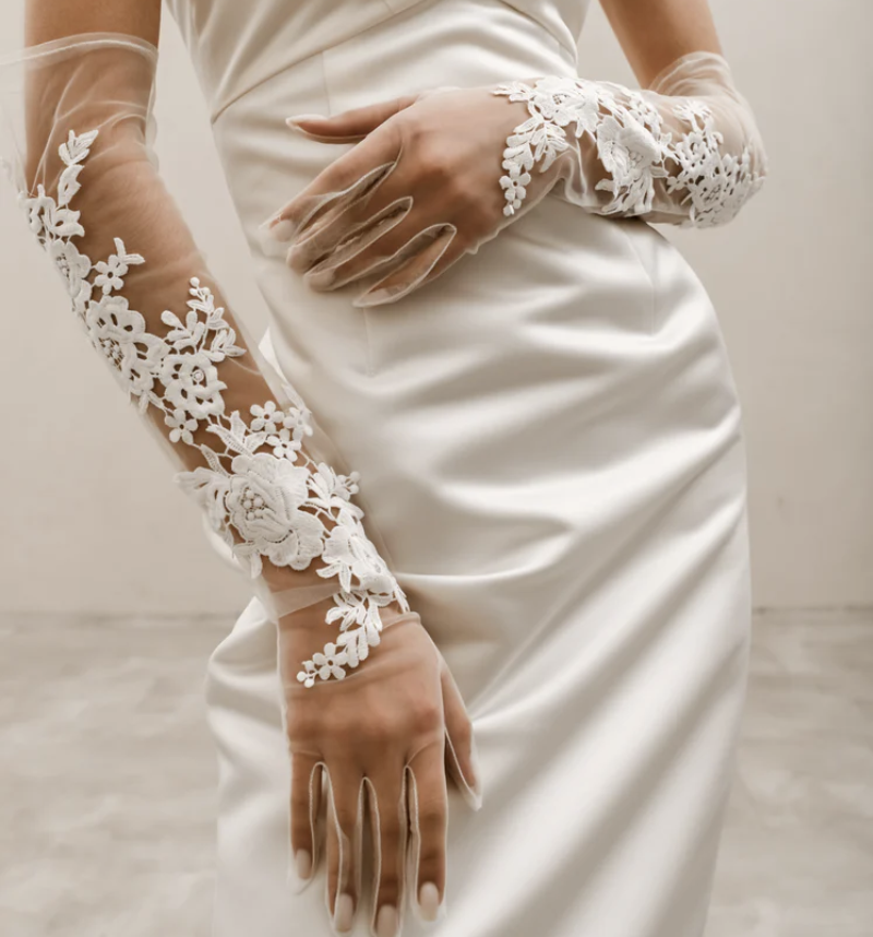 A close-up shows sheer bridal gloves with floral lace appliqués draped over a bride’s arms and hands.