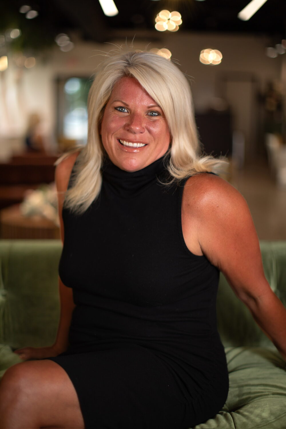 Smiling woman with long silver-blonde hair wearing a sleeveless black dress, seated in a softly lit indoor space.
