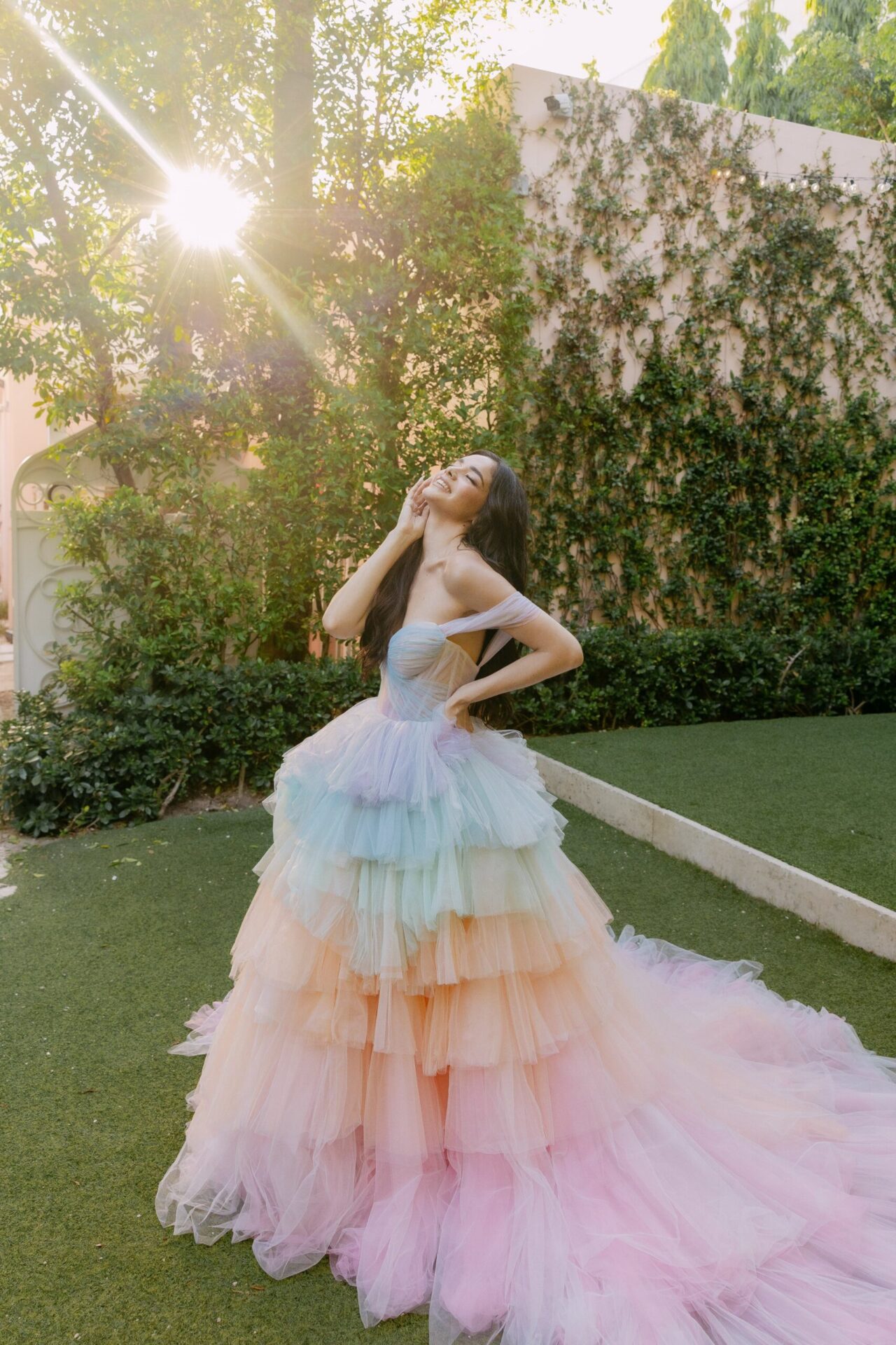 Woman in a tiered pastel rainbow tulle gown standing outdoors with sunlight filtering through trees.