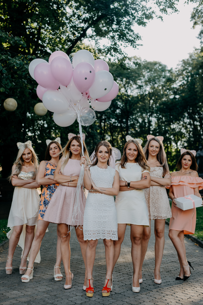 A bride and her bridesmaids wearing light-colored dresses stand together outdoors, holding pastel balloons.