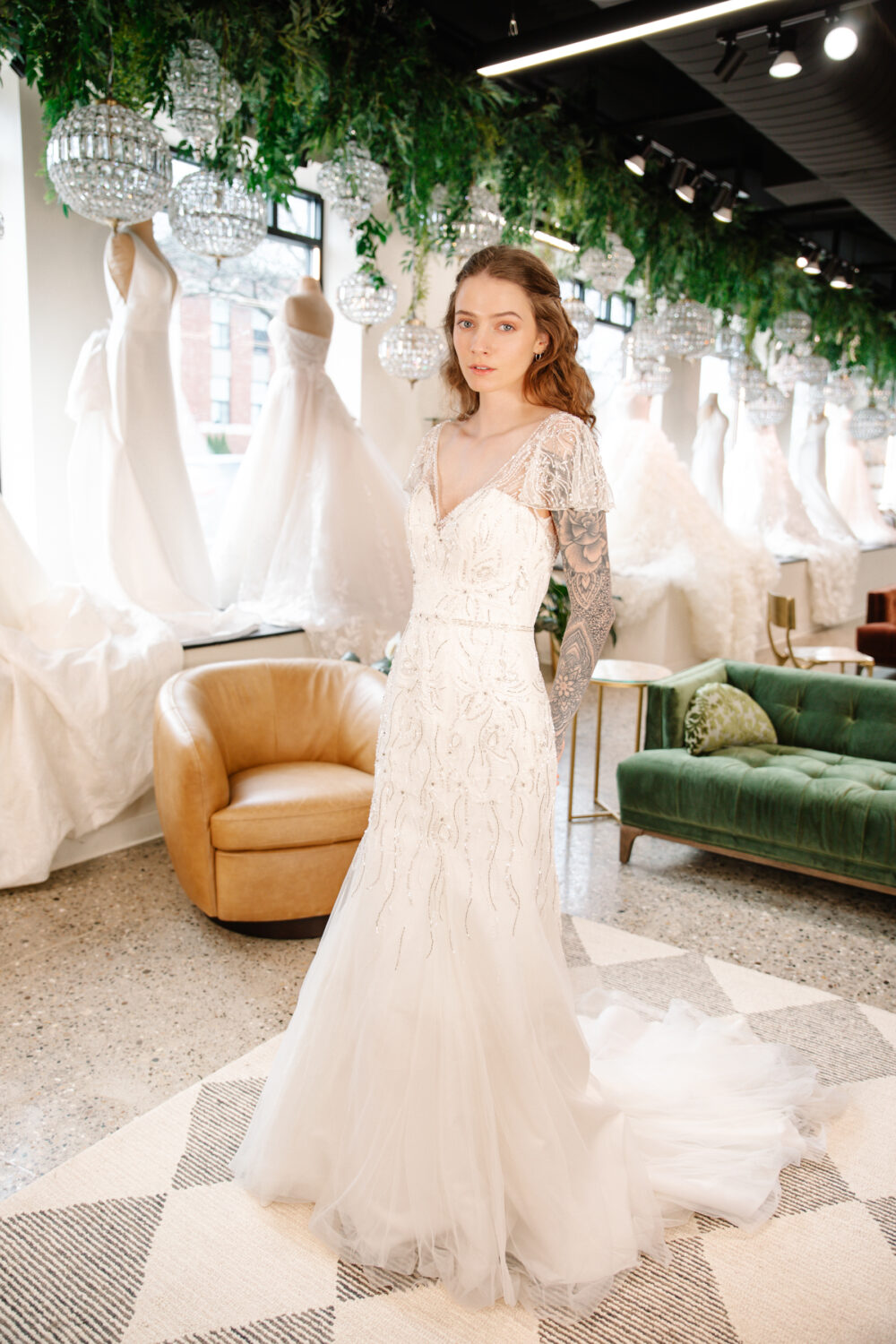 A bride with tattoos wears a fitted beaded wedding dress inside a boutique with gowns displayed behind her.