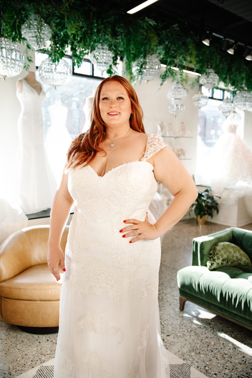 A bride in a lace wedding dress smiles inside a bridal shop with hanging greenery and sparkling chandeliers overhead.