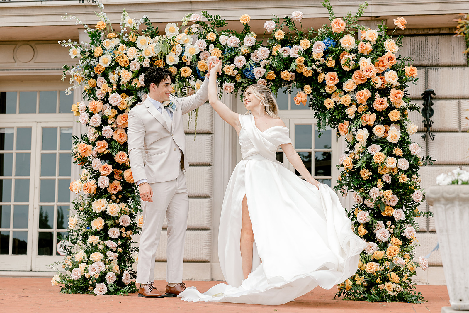 A newlywed couple dances in front of a building decorated with a large arch of peach and white flowers.