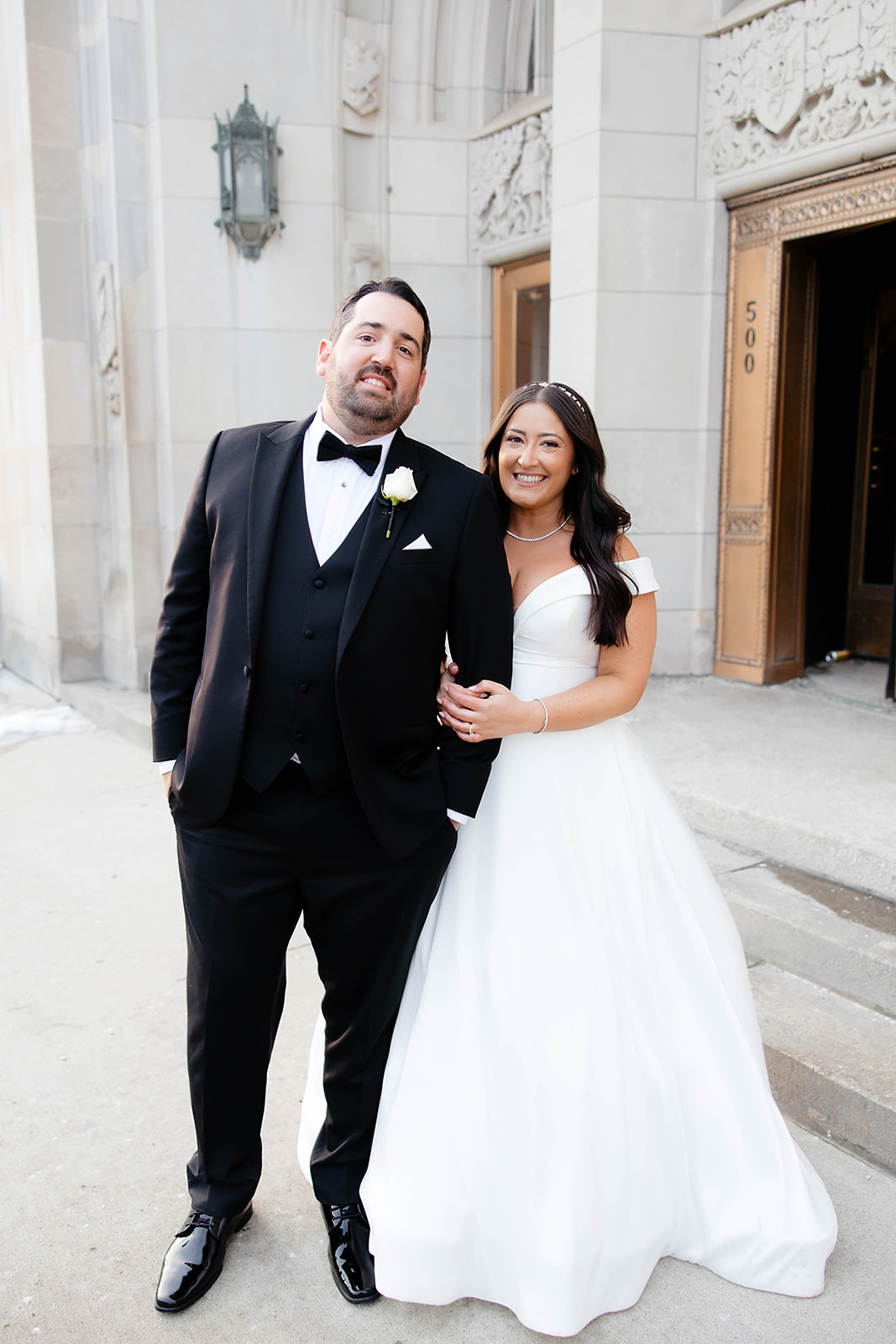 A bride in a full white gown and a groom in a black tuxedo stand together, smiling in front of an ornate stone building.