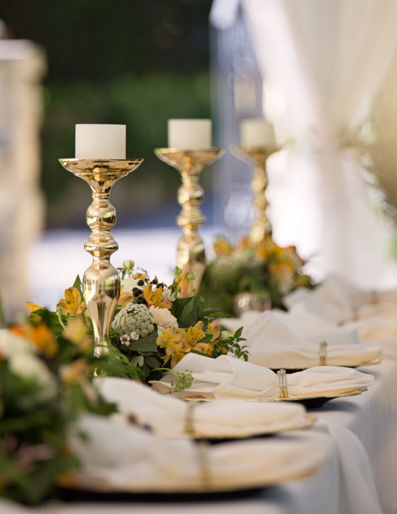 close-up of a wedding reception table shows gold candlesticks, greenery, and neatly arranged plates.