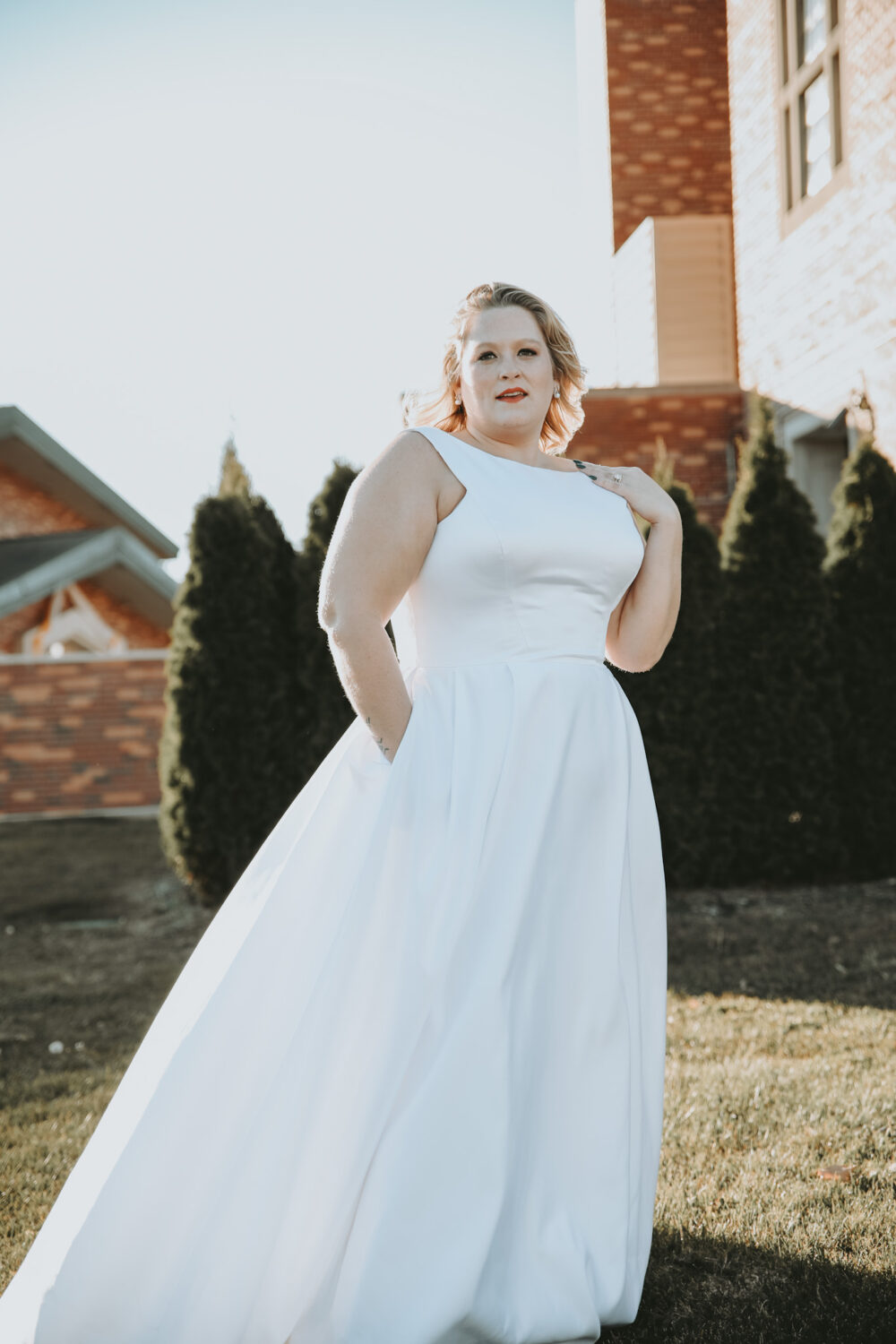 A bride in a simple white sleeveless wedding dress poses outside near a brick building and evergreen trees.