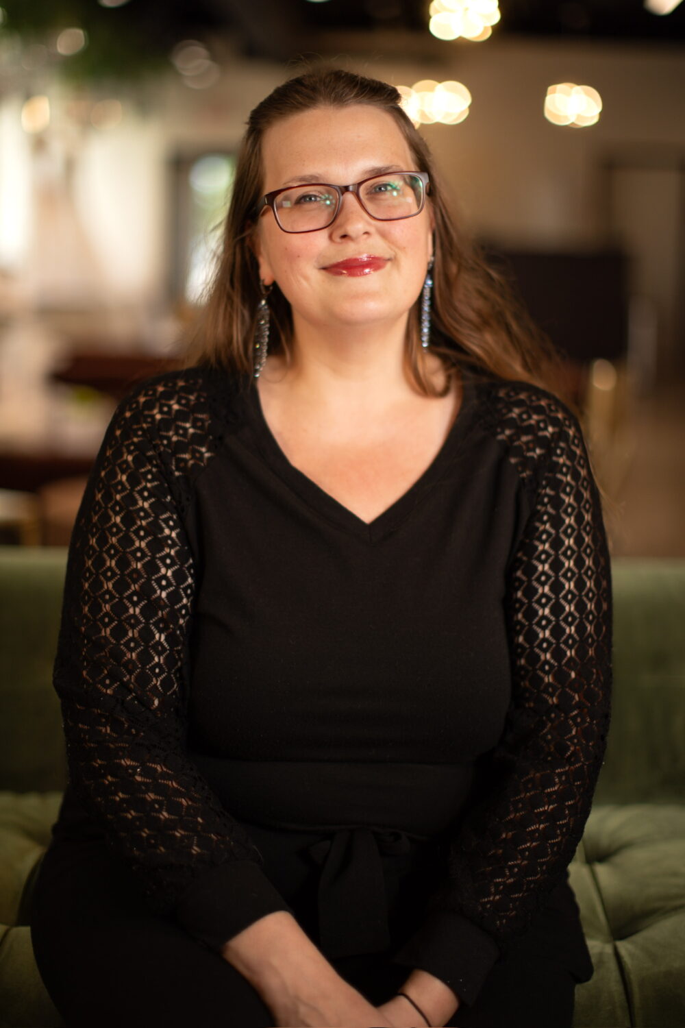 Smiling woman with dark hair and glasses wearing a black lace-sleeve top, seated against a warm, softly blurred indoor background.