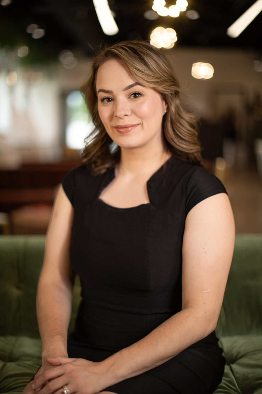 Smiling woman with light brown wavy hair wearing a black sleeveless dress, seated in a warmly lit indoor setting.