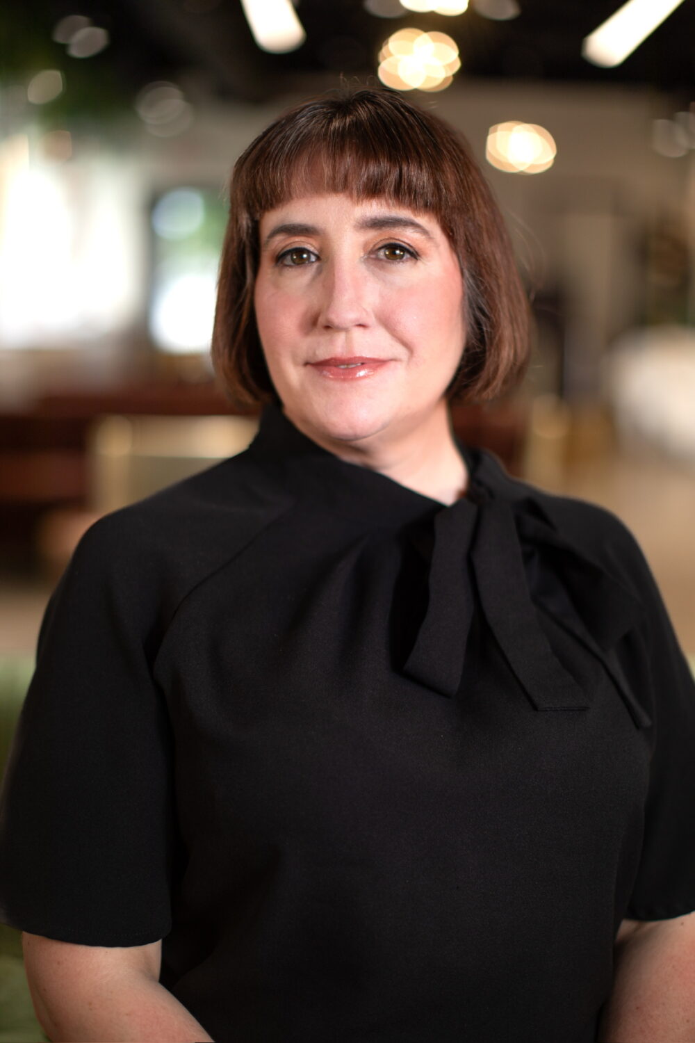 Woman with short dark hair and bangs wearing a black blouse with a bow detail, photographed against a softly blurred indoor background.