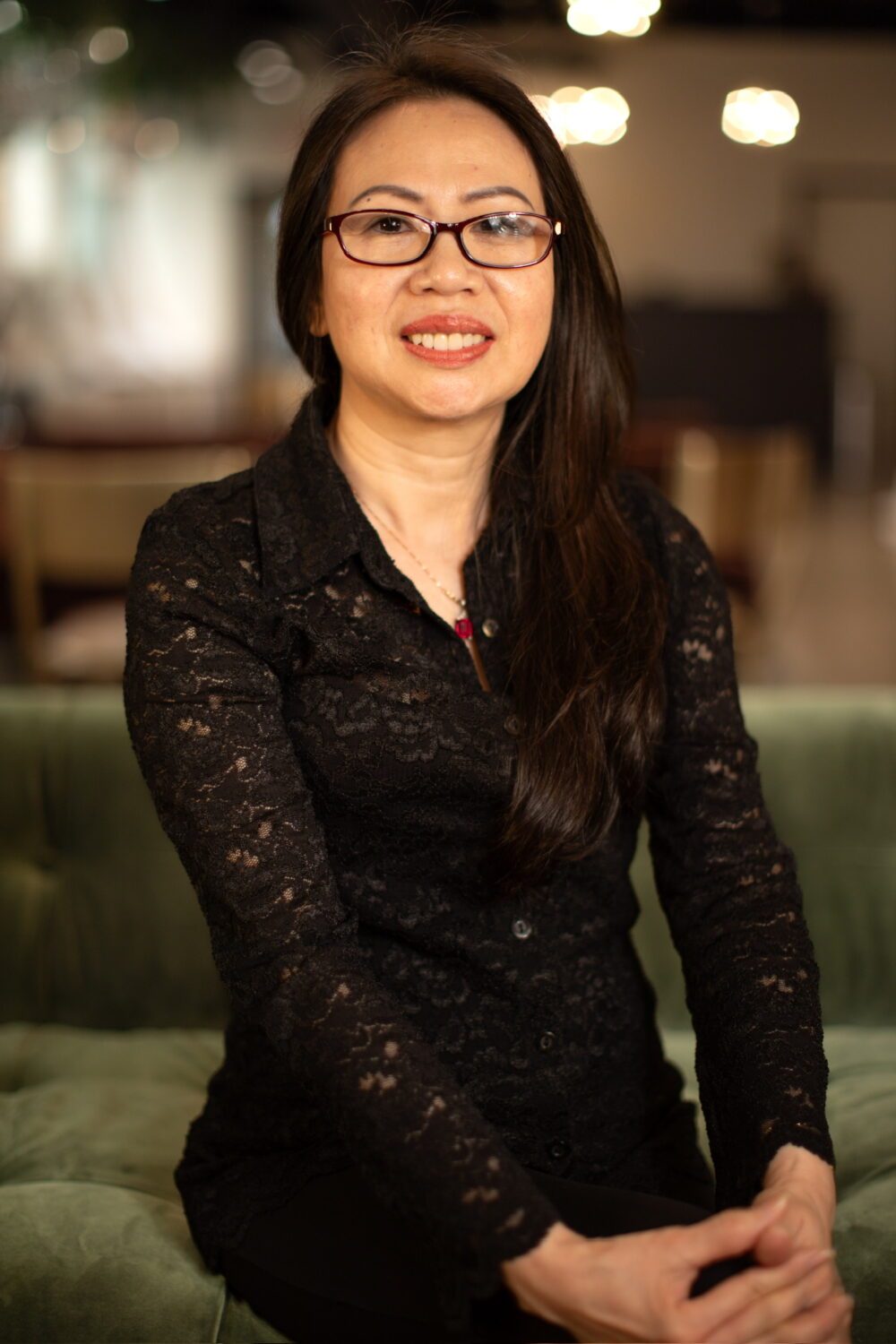 Smiling woman with long dark hair and glasses wearing a black lace top, seated in a warm, softly lit indoor space.