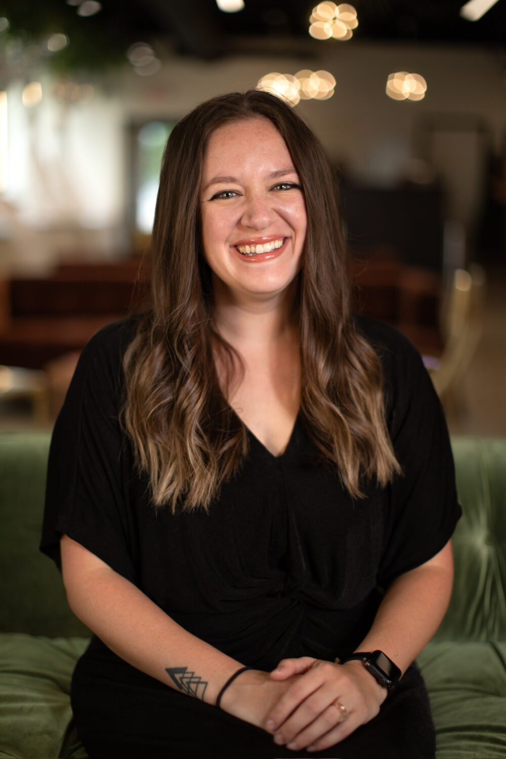 Smiling woman with long brown hair wearing a black short-sleeve top, seated with hands folded in a softly blurred indoor setting.