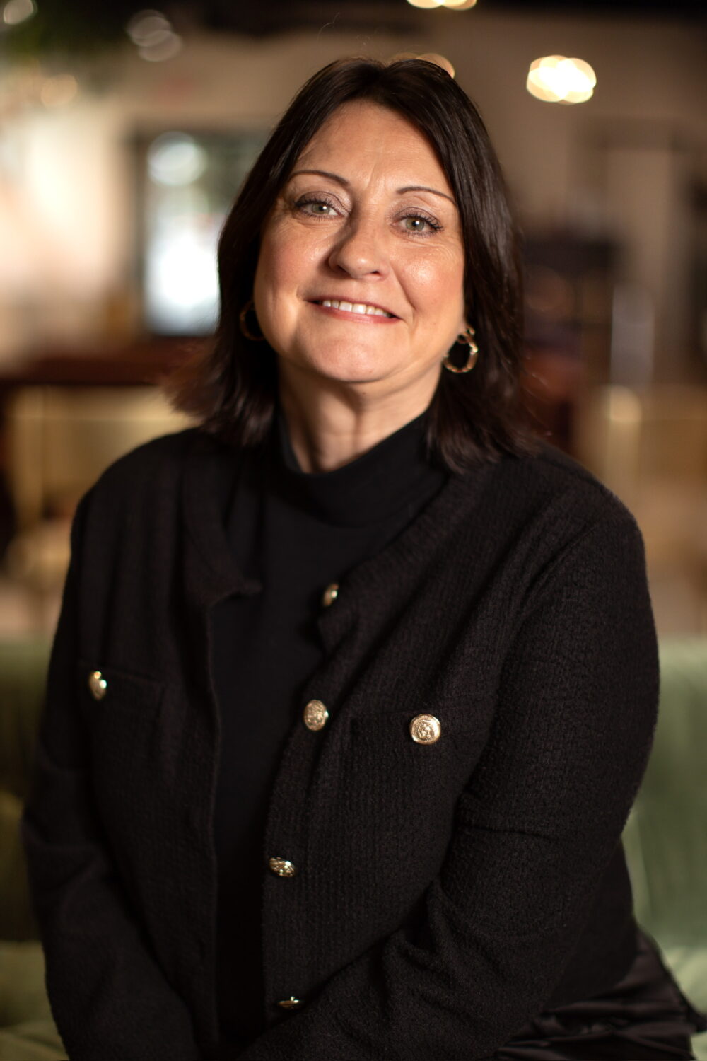 Smiling woman with dark shoulder-length hair wearing a black jacket with gold buttons, photographed in warm indoor lighting.