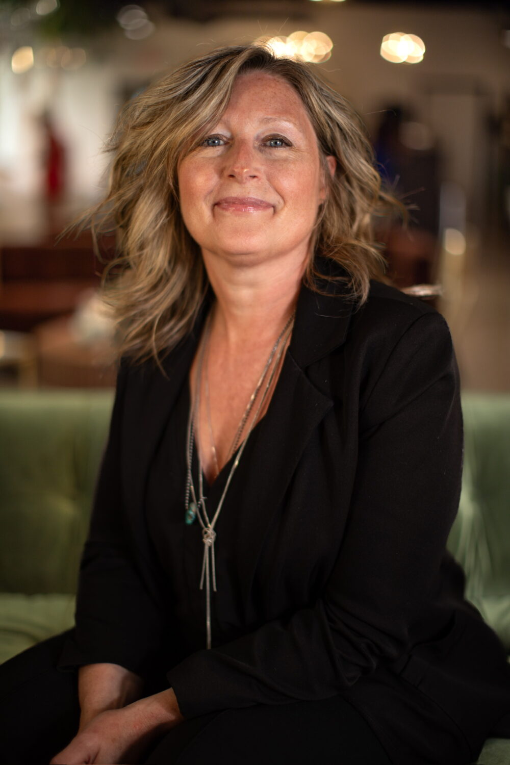 Smiling woman with shoulder-length blonde hair wearing a black blazer and layered necklaces, seated in a warmly lit indoor setting.