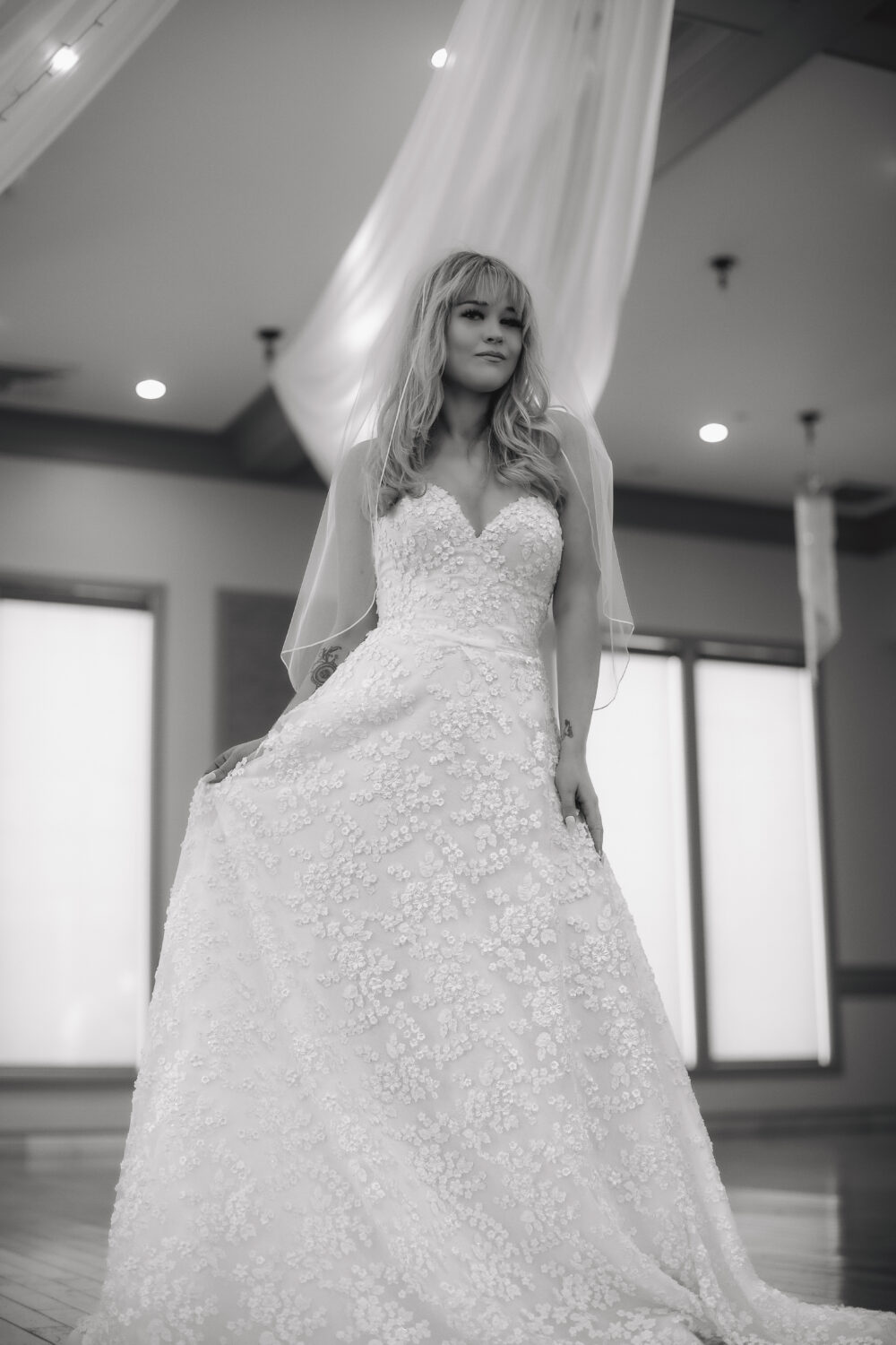 A black-and-white photo shows a bride wearing a strapless lace wedding gown and a veil in a room with draped fabric.