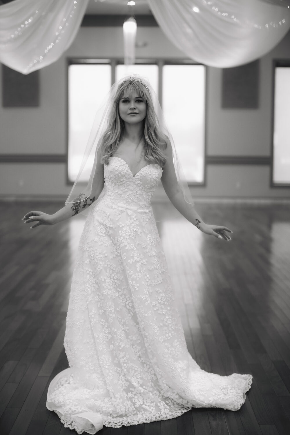 A black-and-white photo shows a bride standing in a lace wedding gown and veil with her arms slightly outstretched.