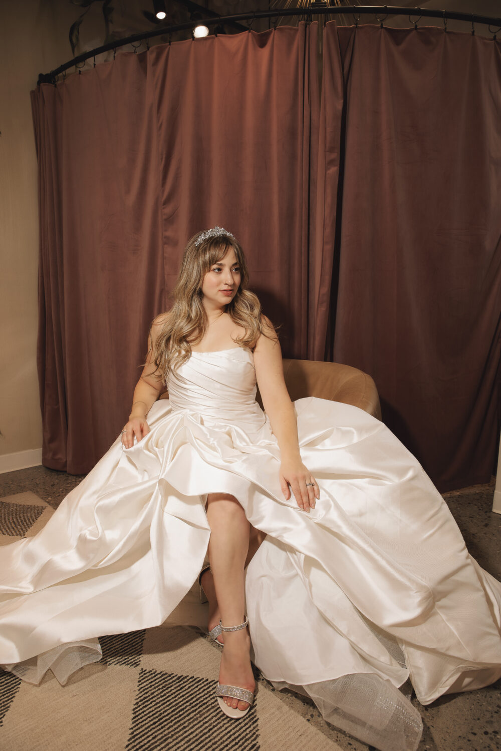 A bride in a satin wedding gown sits in a chair and looks to the side in front of a brown curtain backdrop.