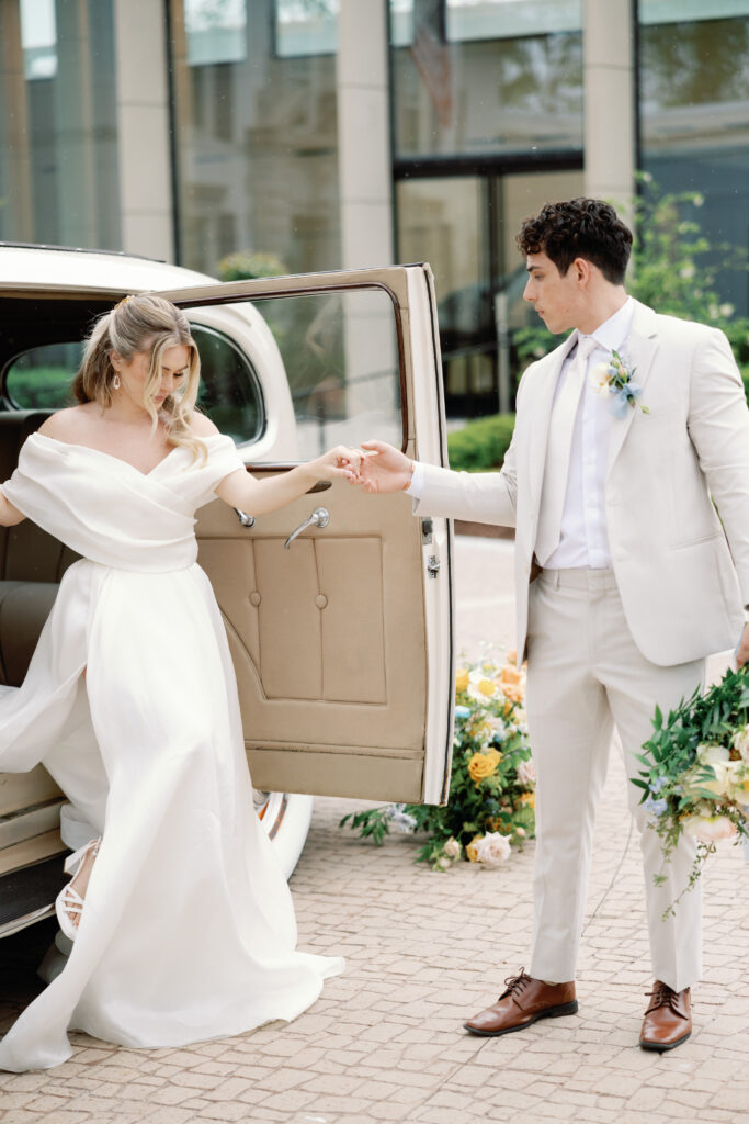 A bride in a white dress steps out of a vintage car while smiling at the groom standing beside her.
