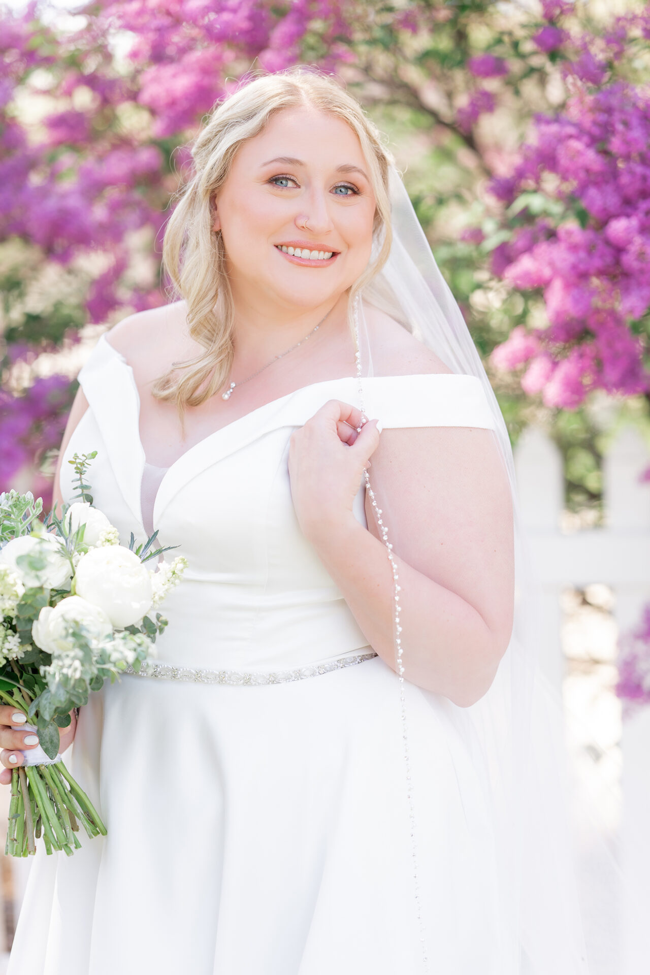 A bride in a white dress smiles while holding a bouquet of white flowers, standing in front of bright pink blossoms.