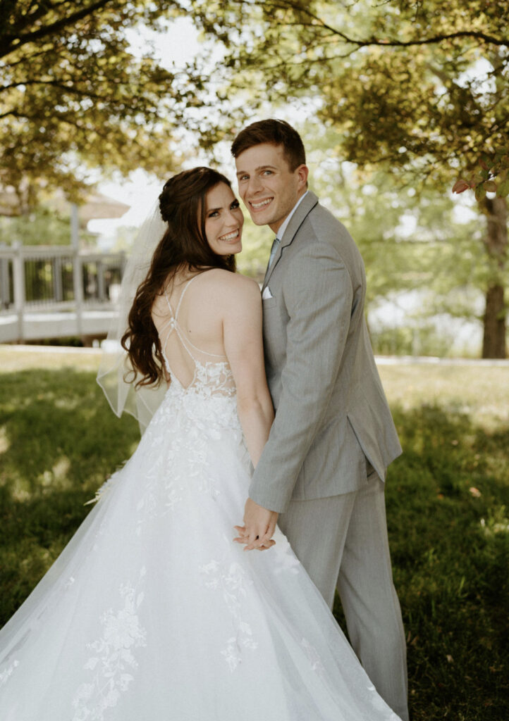 A bride in a lace gown and a groom in a light-colored suit smile and hold each other outdoors beneath a shady tree.