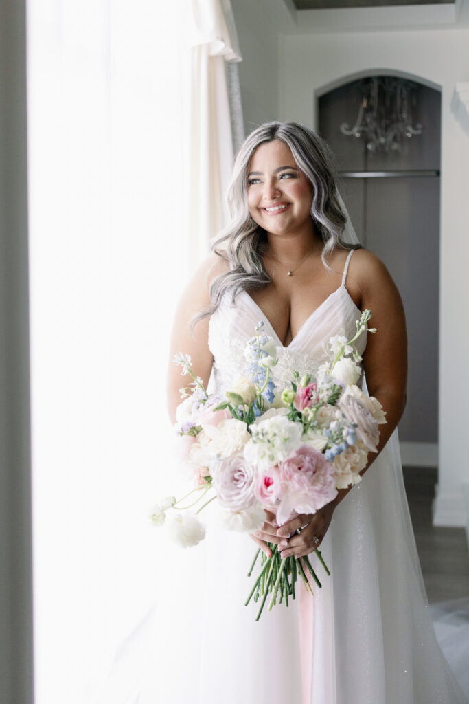 A bride in a sleeveless gown stands by a window holding a bouquet of pale pink and white flowers.