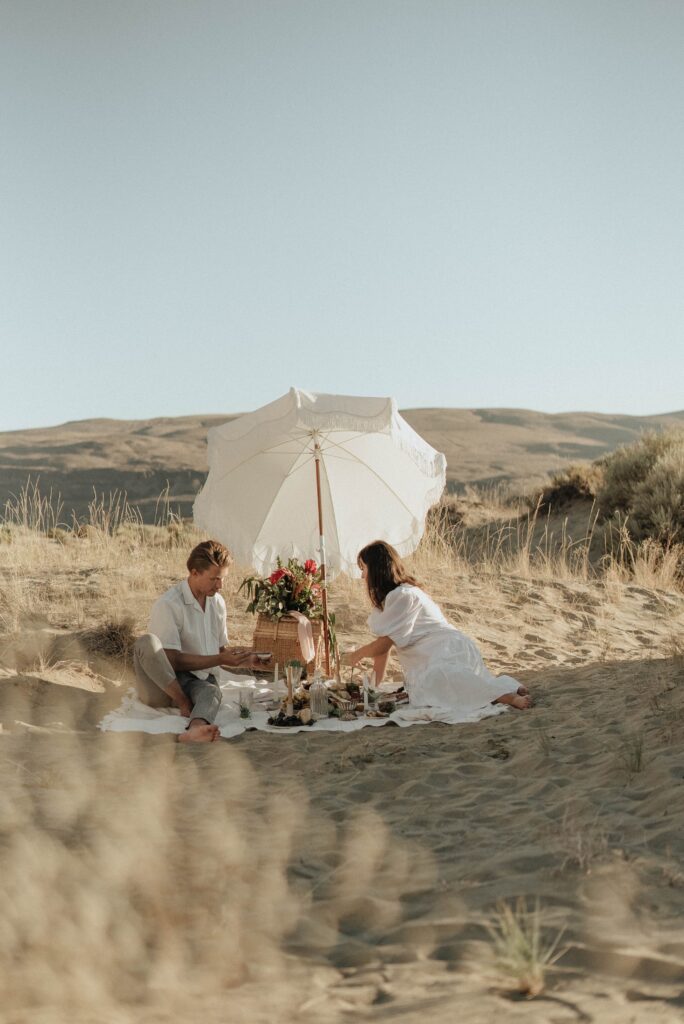A couple sits under a white umbrella on a sandy beach, sharing a picnic surrounded by dunes.