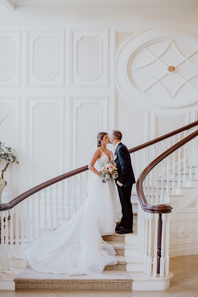 A bride in a long wedding gown and a groom stand together on an elegant curved staircase in a white interior.