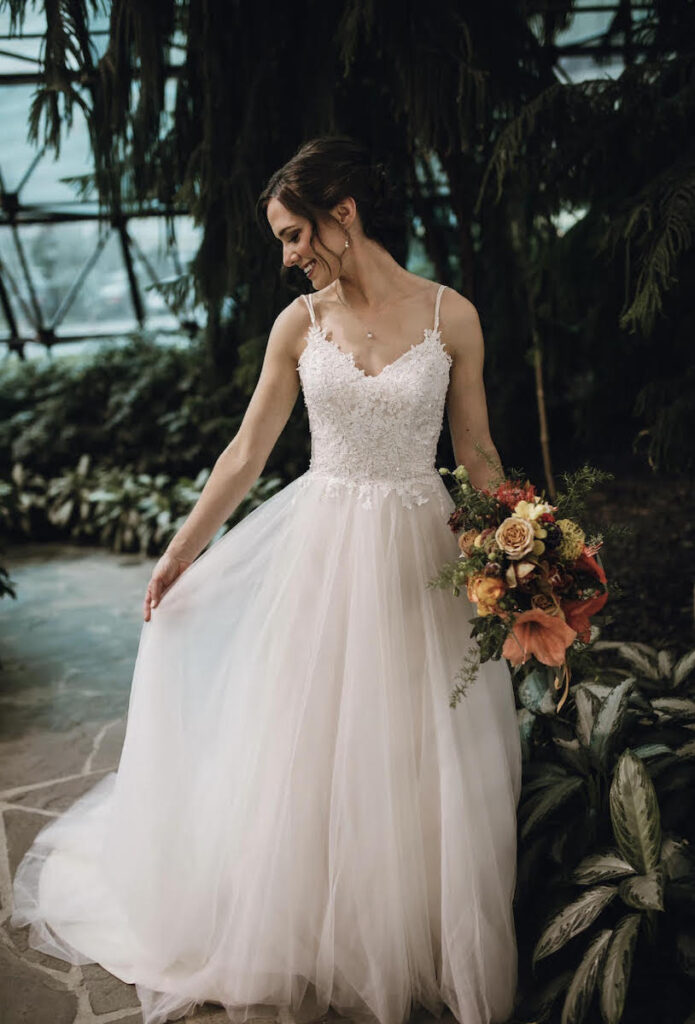Bride standing outdoors in a romantic lace gown, smiling while the wind moves the skirt of her dress.