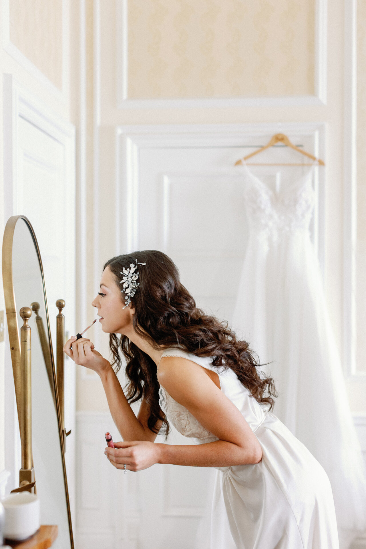 Bride in a white wedding dress applying lipstick while looking into a standing mirror in a softly lit bridal room.